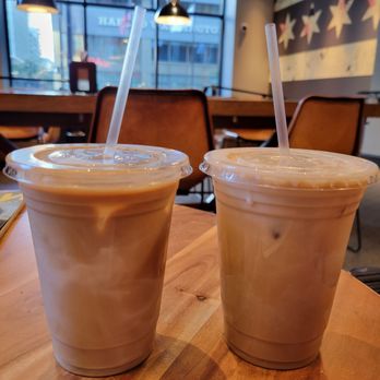 Two Zero Three - Two iced coffee beverages in plastic cups with straws are placed on a wooden table inside a cafe with large windows and modern decor. - Coffee & Tea Near Me - Chicago, Illinois