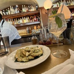 Crispy Zucchini Chips - A cocktail garnished with mint leaves and a straw is placed on a bar counter, alongside a plate of fried vegetables, with a background of liquor bottles and bar equipment.