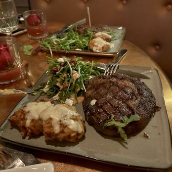 Prohibition - A rectangular plate holds a grilled steak with grill marks, a small side salad with greens and crumbles, and two pieces of mashed potatoes topped with gravy. The plate is on a wooden table with glasses of water, utensils, and a napkin nearby, indicating a restaurant setting. - Cocktail Bars Near Me - Murray, Utah