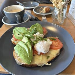 Full Eggs - A gray plate holds avocado toast topped with sliced avocado, poached eggs, tomato slices, and chopped greens. In the background, there is a cup of coffee, a small pastry on a plate, and a vase with dried flowers on a wooden table. The setting appears to be a café with a casual dining atmosphere.