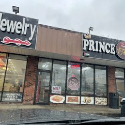 the front of the store - The photo depicts the storefront of Prince Sweets in Bridgeview, IL, featuring signage for jewelry and Middle Eastern sweets. The storefront has large glass windows displaying advertisements for jewelry, gold, diamonds, and various baked goods. There are neon signs indicating the store is open and a 'Help Wanted' sign on the door. The building has a brick facade, and part of a white vehicle is visible on the right side of the image.