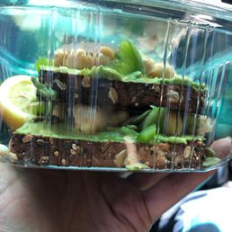 Avocado Toast - A clear plastic container contains avocado toast topped with chickpeas, chopped green peppers, feta cheese, and pumpkin seeds, with a lemon wedge visible.