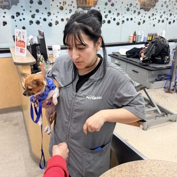 PetSmart - Our Khloe's ready. - A woman in a PetSmart uniform holds a small dog with a blue bandana at the checkout counter. - Pet Training Near Me - Mesa, Arizona
