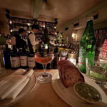 Maison Premiere - Manhattan, bread - The image shows a bar counter with a cocktail in a coupe glass, a plate of sliced bread with a green dip, a large bottle of sparkling water, and a glass of water with ice. In the background, bar staff in white shirts and vests are preparing drinks, with shelves of bottles and bar equipment visible. The setting appears to be a dimly lit, cozy bar or restaurant interior. - Seafood Near Me - Brooklyn, New York