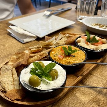 Manouche - Ouche Dipper - Labneh, zesty feta, hummus - A wooden serving board holds three bowls of Mediterranean dips garnished with herbs, accompanied by pita chips, on a restaurant table with glasses and plates. - Salad Near Me - Bloomfield, New Jersey