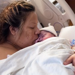 The Tidal Doula - Image: A birthing person kissing their newborn on the face as they lay together in a hospital bed skin to skin. A blanket covers them both.