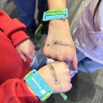 Playdium - ziplining ready - Two children are shown with their fists close together, each wearing a blue wristband with green accents and the word 'PLAUDIUM' visible. They also have temporary tattoos on their hands, which appear to be stamped or printed. The background suggests an indoor setting with a dark, textured floor, and other people are partially visible. The children are dressed casually, one in a red hoodie and the other in a light-colored shirt. - Arcades Near Me - Whitby, Ontario
