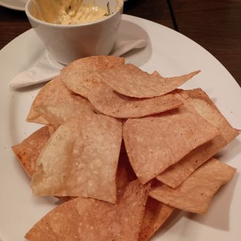 Fork'd - Chips & Queso - A white oval plate contains a serving of crispy, golden-brown tortilla chips. A small white cup of queso dip, with visible herbs, is placed on the plate. The plate is on a dark wooden table, with a napkin underneath the cup. In the background, there is a white plate with a spoon, indicating a dining environment. - Burgers Near Me - Minnetonka, Minnesota