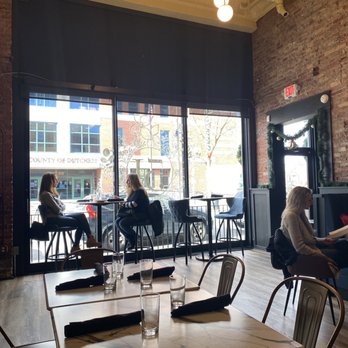 Refinery 51 - Dining area - The image shows the interior of a cafe with a high ceiling, brick walls, and a large windowed front. Three women are seated, two inside near the window and one outside on a high stool. The cafe has a chandelier with multiple round bulbs hanging from the ceiling, and a decorative mirror with greenery on the wall. The table in the foreground has glasses and napkins, indicating a dining setting. - Cocktail Bars Near Me - Poughkeepsie, New York