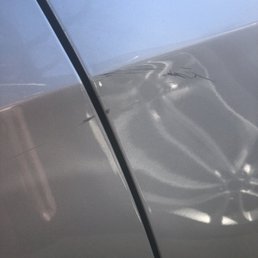 The image depicts a silver car's side panel with a noticeable dent and multiple scratches around the area. The surface reflects light, highlighting the damage, and the photo appears to be taken at close range, focusing on the affected section of the vehicle.