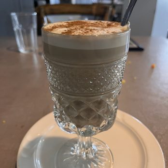 Tiesto's Restaurant - Cappuccino - A layered coffee drink with foam and cocoa powder topping is presented in a textured, decorative glass placed on a white plate. A metal spoon is inserted into the drink, and the background shows a casual dining setting with chairs, tables, and a glass of water. The image emphasizes the presentation and details of the beverage. - Latin American Near Me - Patchogue, New York