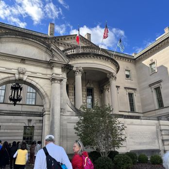 DC Design Tours - Anderson House - The image shows a group of people gathered outside Anderson House, a historic building featuring classical architectural elements such as columns, arches, and decorative stonework. The building has flags flying on its roof, and the sky is partly cloudy with blue and white clouds. The scene appears to be in Washington, DC, and the visitors are likely participating in a guided or self-guided tour of the historic site. - Architectural Tours Near Me