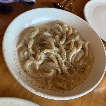 PASTA - Caccio e Pepe perfection - A ceramic bowl contains cooked noodles in broth, topped with black pepper, on a wooden table with utensils and plates nearby. - Italian Near Me - Miami, Florida