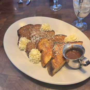 The Henry - Cinnamon Sugar French Toast - French toast topped with whipped cream, powdered sugar, and served with syrup on a white plate, accompanied by drinks on a wooden table. - New American Near Me - Phoenix, Arizona