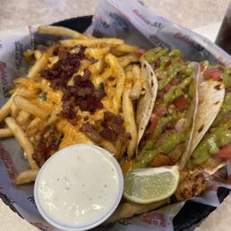 Grilled Shrimp Tacos - A basket contains two tacos with grilled chicken, diced tomatoes, and green sauce, accompanied by French fries topped with shredded cheese and bacon bits. A lime wedge and a small cup of creamy white sauce are also included. The basket is lined with branded paper from Bubba's 33, and the setting appears to be a casual dining restaurant.