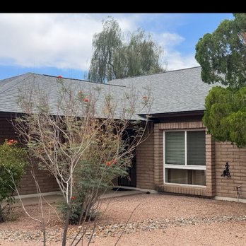 Tip Top Roofing Service - New roof - A brick house with a gray shingled roof, a large window, and a front yard with trees and bushes, under a partly cloudy sky. - Roofing Near Me - Scottsdale, Arizona