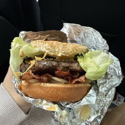 Bacon Cheeseburger - A bacon cheeseburger featuring lettuce, a beef patty, melted cheese, bacon strips, and a sesame seed bun is held in a person's hand. The burger is wrapped in crumpled aluminum foil, with some foil and condiments visible. The background shows the interior of a vehicle, with a dark seat and part of a beige sleeve visible.