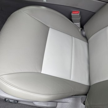 Cibola Upholstery - The image shows a car seat with leather upholstery, combining light gray and white sections. The seat has visible stitching along the edges and a seatbelt buckle positioned at the side. The surrounding interior includes a dark carpeted floor and part of the vehicle's side panel. The seat appears clean and well-maintained. - Furniture Reupholstery Near Me - Corrales, New Mexico