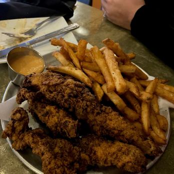 Original Gravity - Chicken strips w/fries - A metal plate holds fried chicken tenders, French fries, and a small container of dipping sauce, on a restaurant table. - New American Near Me - Phoenix, Arizona