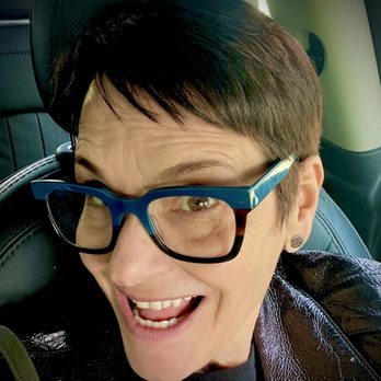 Elixir Salon - A person with short dark hair and large blue glasses is smiling broadly in a selfie. They are wearing earrings and a shiny black jacket. The background indicates they are inside a vehicle, with visible car seats and interior details. The lighting suggests natural light coming from outside the vehicle. - Hair Stylists Near Me - Berkeley, California