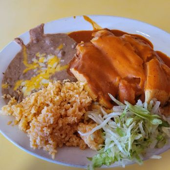 Chalo's - Pork Tamale - A white oval plate contains a Mexican-style meal with a tamale covered in sauce, Mexican rice, refried beans topped with melted cheese, and shredded lettuce. The plate is placed on a yellow table, and part of a wooden surface is visible in the background. - Mexican Near Me - Globe, Arizona