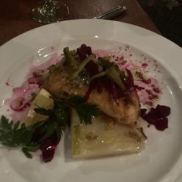 Salmon - A white plate holds a cooked fish fillet topped with herbs and vegetables, including leafy greens and possibly beets or radishes. The dish is garnished with a sprig of parsley and served with a sauce or puree underneath. A glass of white wine and a fork are visible on a wooden table in a dimly lit setting.