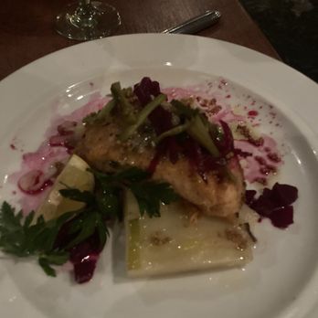 RIS - Salmon - A white plate holds a cooked fish fillet topped with herbs and vegetables, including leafy greens and possibly beets or radishes. The dish is garnished with a sprig of parsley and served with a sauce or puree underneath. A glass of white wine and a fork are visible on a wooden table in a dimly lit setting. - New American Near Me - Washington, DC, District of Columbia