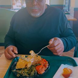 A man with glasses and a black beanie is seated at a table in a restaurant, eating a plate of soul food including greens, fried chicken, and other dishes.