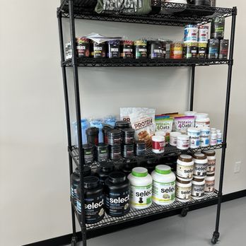 The Smoothie Stop - Protein - The image shows a black metal shelving unit with four shelves, filled with dietary supplements, protein powders, and fitness products. The top shelf contains large containers of supplements, while the second shelf has smaller containers and cans. The third shelf displays various protein powders in jars and pouches, and the bottom shelf holds larger protein containers and bottles. The items are organized and labeled, with some products from brands like Select and others with visible labels. The shelving unit is positioned against a plain wall with a power outlet nearby. - Juice Bars & Smoothies Near Me - Riverview, Florida