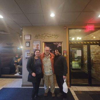 Cool Basil - Three people stand together in front of a restaurant entrance, with a woman on the left, a man in the middle, and another man on the right holding a white bag. The background includes a wall with framed items, a fire extinguisher, and a glass door leading outside. The restaurant interior features a ceiling with recessed lighting and a seating area visible through a window on the left. - Thai Near Me - Clive, Iowa