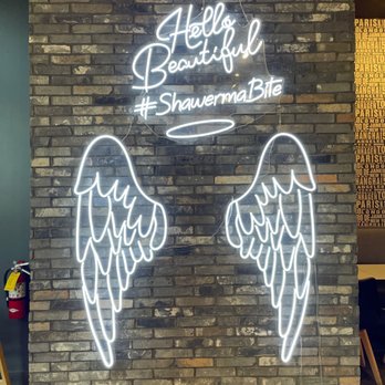Shawerma Bite - A neon sign on a brick wall displays 'Hello Beautiful' with the hashtag '#ShawermaBite' and features angel wings outline in white neon. - Mediterranean Near Me - Columbus, Ohio