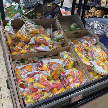 ALDI - Bags of pre-packaged mixed bell peppers in red, yellow, and green are stacked in a cardboard display box at a grocery store. The display is situated on a tiled floor, with other produce items, including a box of limes, visible nearby. The store shelves and other shoppers are partially visible in the background, indicating a typical grocery store setting. - Grocery Near Me - Deerfield Beach, Florida