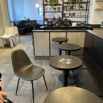 Gregorys Coffee - The image shows the interior of a coffee shop with a seating area that includes a mix of chairs and tables. There are black and brown chairs, some stacked, and small round tables. A black and wood display shelf with potted plants and coffee products separates the seating area from the counter. The background includes a window, pendant lights, and a white wall with shelves. The overall decor is modern with a neutral color palette. - Coffee & Tea Near Me - Washington, DC, District of Columbia