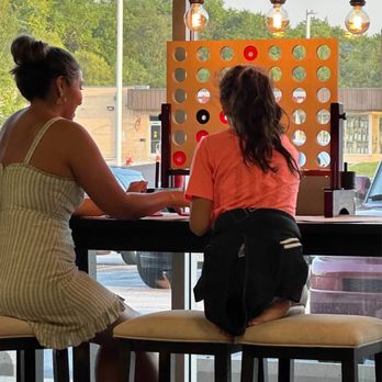 Mastic Gelato - A woman and a child are seated at a cafe table playing Connect Four, with large windows behind them showing trees and parked cars. - Ice Cream & Frozen Yogurt Near Me - Orland Park, Illinois