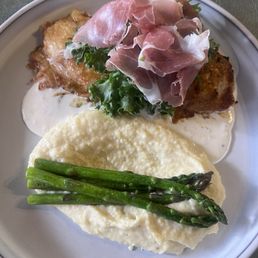 Anabelle Chicken - A dish with mashed potatoes topped with asparagus, a fried item with sauce, and prosciutto on greens, served on a white plate.