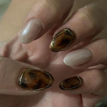 Uni Nails - Perfect one of a kind design by Helen. - The image shows a close-up of a hand with five nails, three of which have a tortoiseshell pattern with brown and black swirls, bordered by a thin gold outline. The remaining two nails are painted in a natural, glossy, light beige color. The nails are well-shaped and appear to be professionally manicured. The background is blurred, focusing attention on the nails and hand. - Nail Salons Near Me - Tempe, Arizona