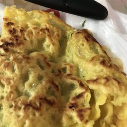 Banh Xeo - A close-up image of a Vietnamese Banh Xeo, a savory pancake with a crispy, golden-brown surface. The pancake appears to contain green vegetables or herbs inside. A black utensil is positioned above the food, and the dish is placed on white paper or napkin. The photo emphasizes the texture and color of the Banh Xeo.