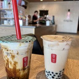 Winter Melon Oolong Tea - Two bubble tea drinks from Gong cha are positioned on a wooden table inside a cafe. The drink on the left has a marbled appearance with brown and white swirls and a red straw, while the drink on the right contains black tapioca pearls at the bottom and has a clear plastic lid. The background shows a cafe counter with staff and menu screens, slightly out of focus.