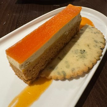 Junction - Tomato Dessert - A rectangular layered dessert with an orange glaze on top, served on a white plate. It is accompanied by a blue cheese cracker and a small amount of orange sauce on the plate. The dessert appears to have a sponge cake base with a creamy middle layer. - New American Near Me - Essex Junction, Vermont