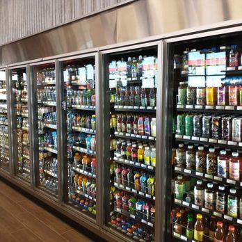 Sheetz - A series of glass-door refrigerated coolers containing a variety of bottled beverages such as juices, sodas, and flavored drinks. The coolers are aligned in a row along a store wall, with reflections visible on the glass doors. The store has a modern interior with wood-look flooring and a metallic panel above the coolers. - Gas Stations Near Me - Columbus, Ohio
