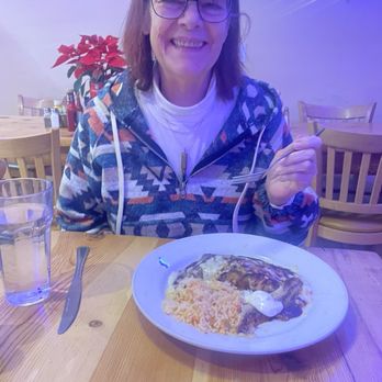 Tune-Up Cafe - My very happy momma - A woman with shoulder-length brown hair and glasses is sitting at a wooden table in a restaurant, smiling at the camera. She is wearing a patterned jacket over a white turtleneck. On the table, there is a plate of food that appears to include rice, a meat dish with sauce, and a dollop of sour cream or similar topping. A glass of water and a knife are also visible on the table. The background shows wooden chairs, a poinsettia plant, and some artwork on the wall, with blue lighting and holiday decorations. - New Mexican Cuisine Near Me - Santa Fe, New Mexico