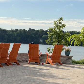 Menomonie Public Library - 2 people sitting and reading in orange Adirondack chairs on the library terrace, facing a calm lake and green trees in the background. - Libraries Near Me - Menomonie, Wisconsin Menomonie Public Library - 2 people sitting and reading in orange Adirondack chairs on the library terrace, facing a calm lake and green trees in the background. - Libraries Near Me - Menomonie, Wisconsin