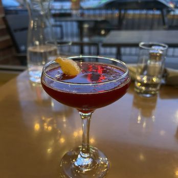 Social Oak Wine Bar - Bite Me - A red-colored cocktail with a lemon wedge garnish is served in a coupe glass on a wooden table inside a wine bar. In the background, there are water glasses, a pitcher, and a window showing parked cars and illuminated signs outside. The setting includes outdoor seating visible through the window, and the lighting suggests evening time. - Wine Bars Near Me - Eagle, Colorado