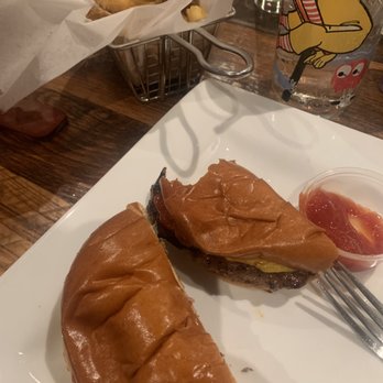 Public Kitchen - Burger.....which was delicious! - Two cheeseburgers with toasted buns and beef patties are placed on a white square plate. One burger has a visible slice of cheese, and a fork is inserted into the other burger. A small cup of ketchup is also on the plate. In the background, there is a basket of fried food, a glass with a cartoon character, and a wooden table surface. - Gastropubs Near Me - Wakefield, Massachusetts