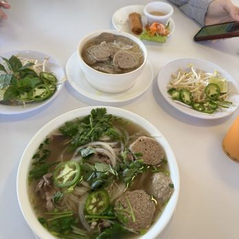 I Luv Pho - The image shows a table set with Vietnamese dishes, including a large bowl of pho with beef, herbs, onions, and sliced jalapeños, and a smaller bowl of beef noodle soup with meatballs. There are also plates of bean sprouts, sliced jalapeños, and fresh herbs, along with spring rolls, a small salad, and a dipping sauce. A person is seated at the table, holding a smartphone, with part of their arm and clothing visible. - Vietnamese Near Me - Irving, Texas