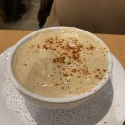 Creamy crab soup - A bowl of creamy crab soup with a thick, smooth consistency, topped with a dusting of spices, is placed on a white plate with a decorative paper doily underneath. The background shows part of a person's beige shirt and a wooden table surface, indicating a dining setting.
