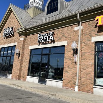 Faces by Freda - The image shows the storefront of Faces by Freda, a beauty salon situated in a brick building with large glass windows and doors. The signage displays the salon's name prominently above the entrance. The building features exterior lighting fixtures and a sidewalk in front. Adjacent signage indicates other businesses or services within the same complex. The scene is captured during daylight with a clear blue sky. - Waxing Near Me - Novi, Michigan