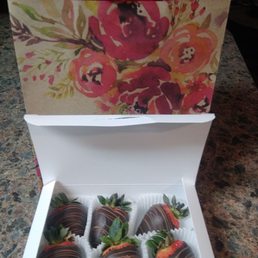 a box of chocolate covered strawberries - A white box containing six strawberries dipped in dark chocolate with drizzled white chocolate is placed on a speckled granite surface. Each strawberry has green leaves attached. Behind the box, there are two shopping bags, one with a floral watercolor design and the other in solid red, on a dark background.