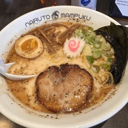 Black Tonkotsu - A bowl of ramen featuring pork slices, half a soft-boiled egg, chopped green onions, seaweed, and narutomaki, served in a restaurant setting.