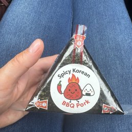 BBQ pork - A triangular onigiri rice ball with spicy Korean BBQ pork filling is held in a person's hand, with denim jeans visible in the background.